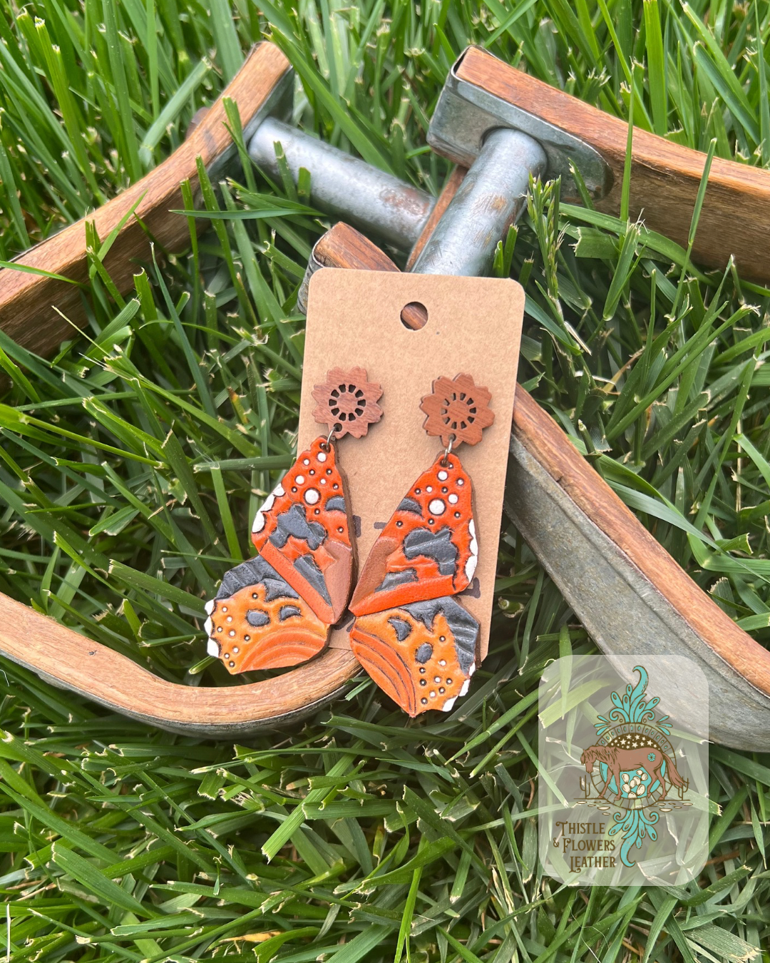 Tooled and painted leather earrings shaped and painted like painted lady butterfly wings. Paint is various shades of oranges, black, and white. Earrings have wooden stud piece. Photographed on wooden and metal stirrups on grass. Logo is in bottom right corner of photo.