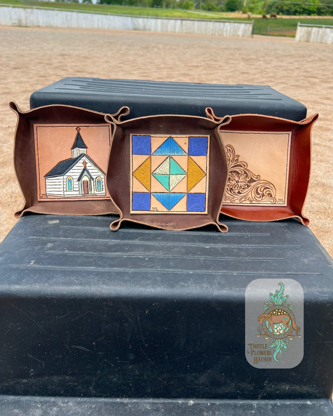 3 valet trays on mounting block in arena. Left valet tray has a tooled and painted church on the base, center tray has a tooled and painted quilt square on the base, and the right valet tray has a traditional western floral design. Logo is in bottom right of photo.