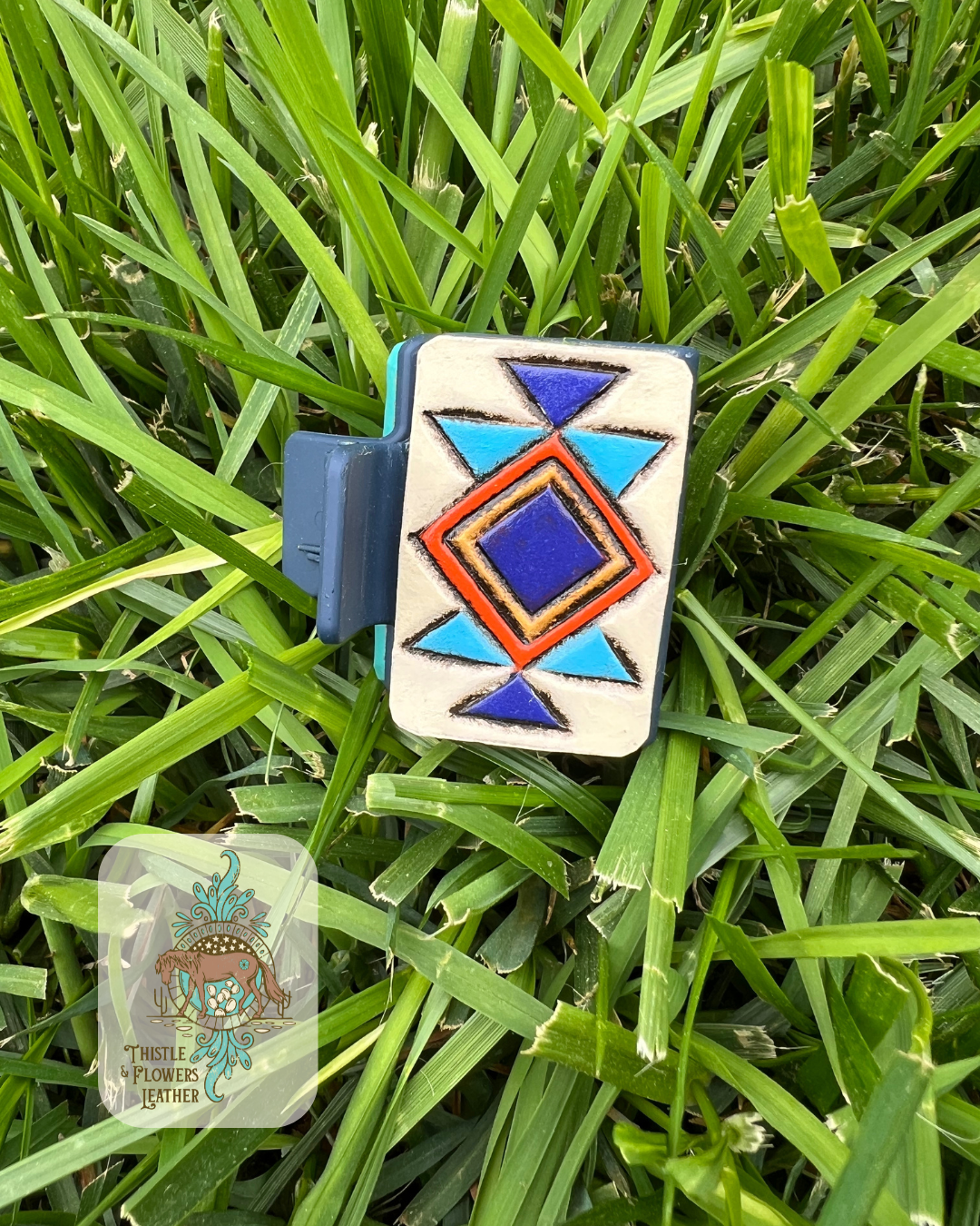 Navy blue base Hairclip with tooled and painted leather patches. Patches has a southwestern design tooled and painted on them using cream, dark blue, light blue, orange, and yellow colors. Photographed on grass with logo in bottom left of photo.