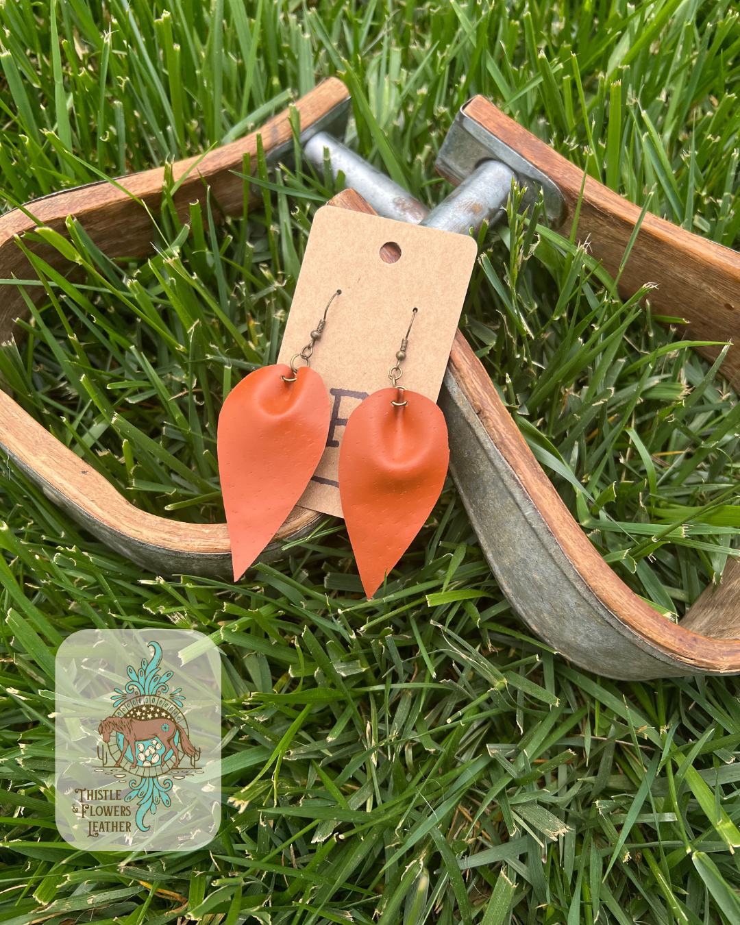 Smooth light brown reverse teardrop leather earrings. Photographed on wooden and metal stirrups on grass with logo on bottom left corner of photo.
