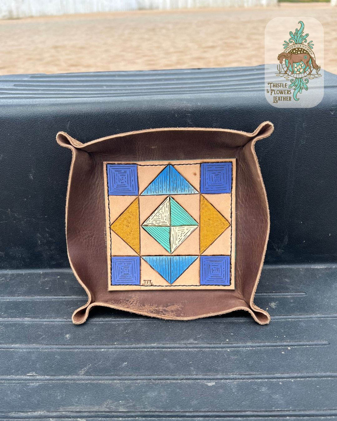 Valet tray with a tooled and painted leather base. Base is tooled and painted to appear like a barn quilt or quilt square. Painted colors are dark blue, light blue, yellow, white, & turquoise. Tray is on a mounting block step. Logo is in top right corner of photo.