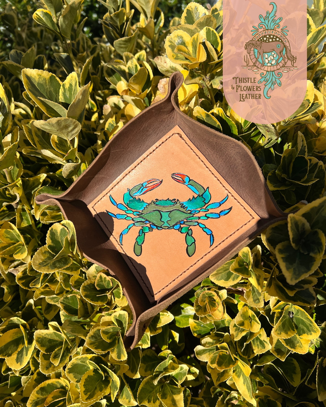 Leather Valet tray with painted blue crab on bottom of tray. Tray is propped up in a green and yellow bush, with strong golden hour sunlight highlighting colors. Photo is watermarked with the Thistle & Flowers Leather logo.