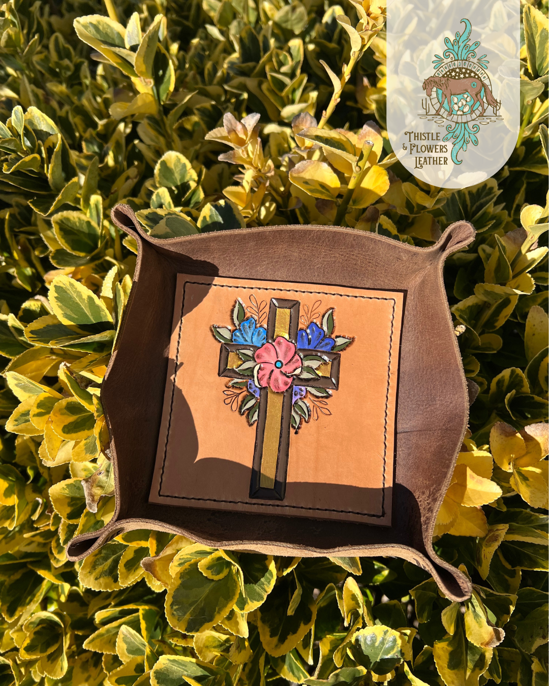 Leather Valet tray with tooled and painted cross with flowers around it on bottom of tray. Tray is propped up in a green and yellow bush, with strong golden hour sunlight highlighting colors and metallic paint in cross. Photo is watermarked with the Thistle & Flowers Leather logo.