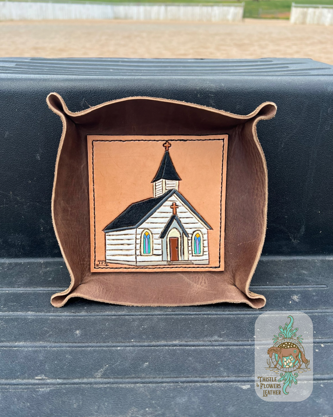 Brown leather valet tray with a hand tooled and painted leather base. Base has a white chapel with stained glass windows, brown door, and black roof tooled and painted on it. Tray is sitting on mounting block. Logo is in bottom right of photo.