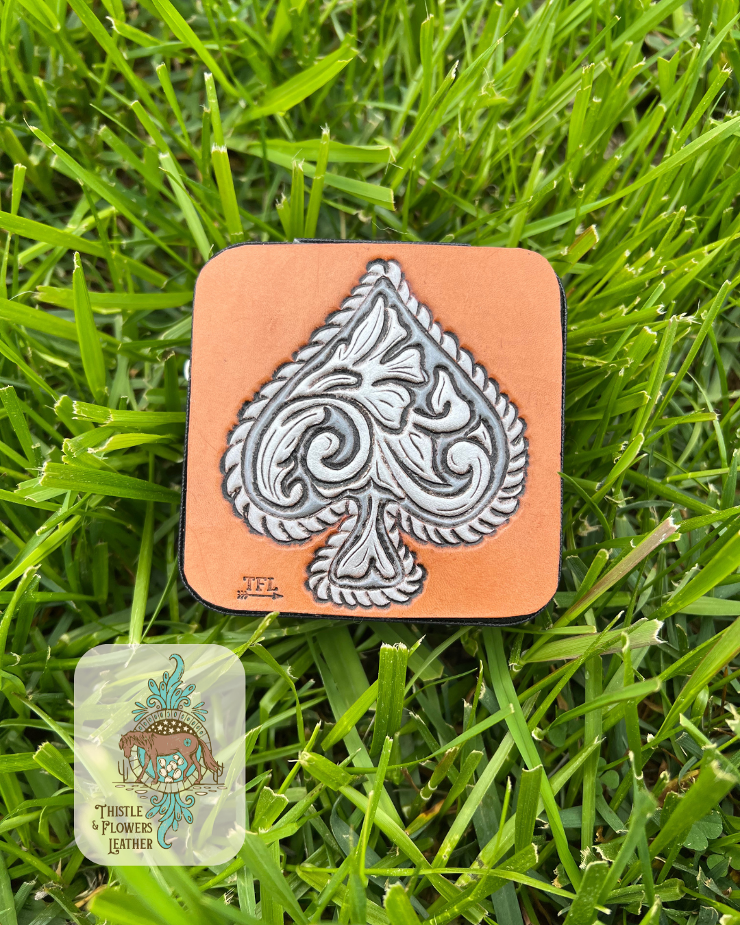 Mini travel jewelry box with a tooled and painted leather patch. Patch features a silver concho looking spade. Spade is painted in various shades of metallic silver. Jewelry box is on grass. Logo is in bottom left of photo.