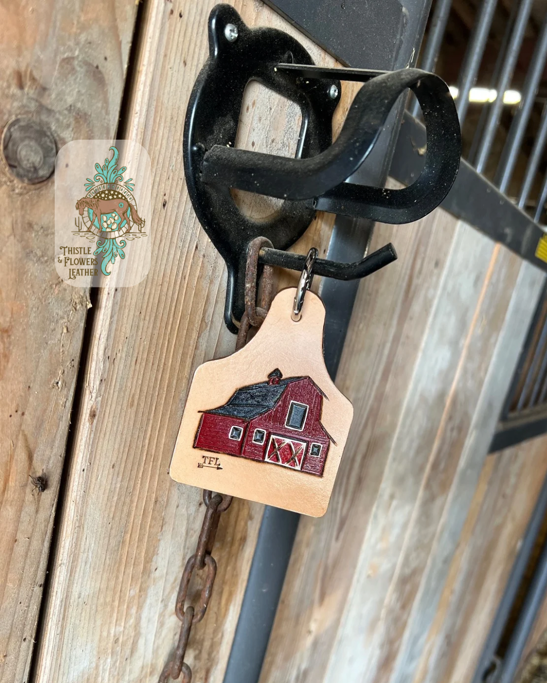 Cowtag shaped tooled leather keychain with a tooled and painted red barn on on it. Keychain is hanging on a stall door. Logo is on the top right of the photo.