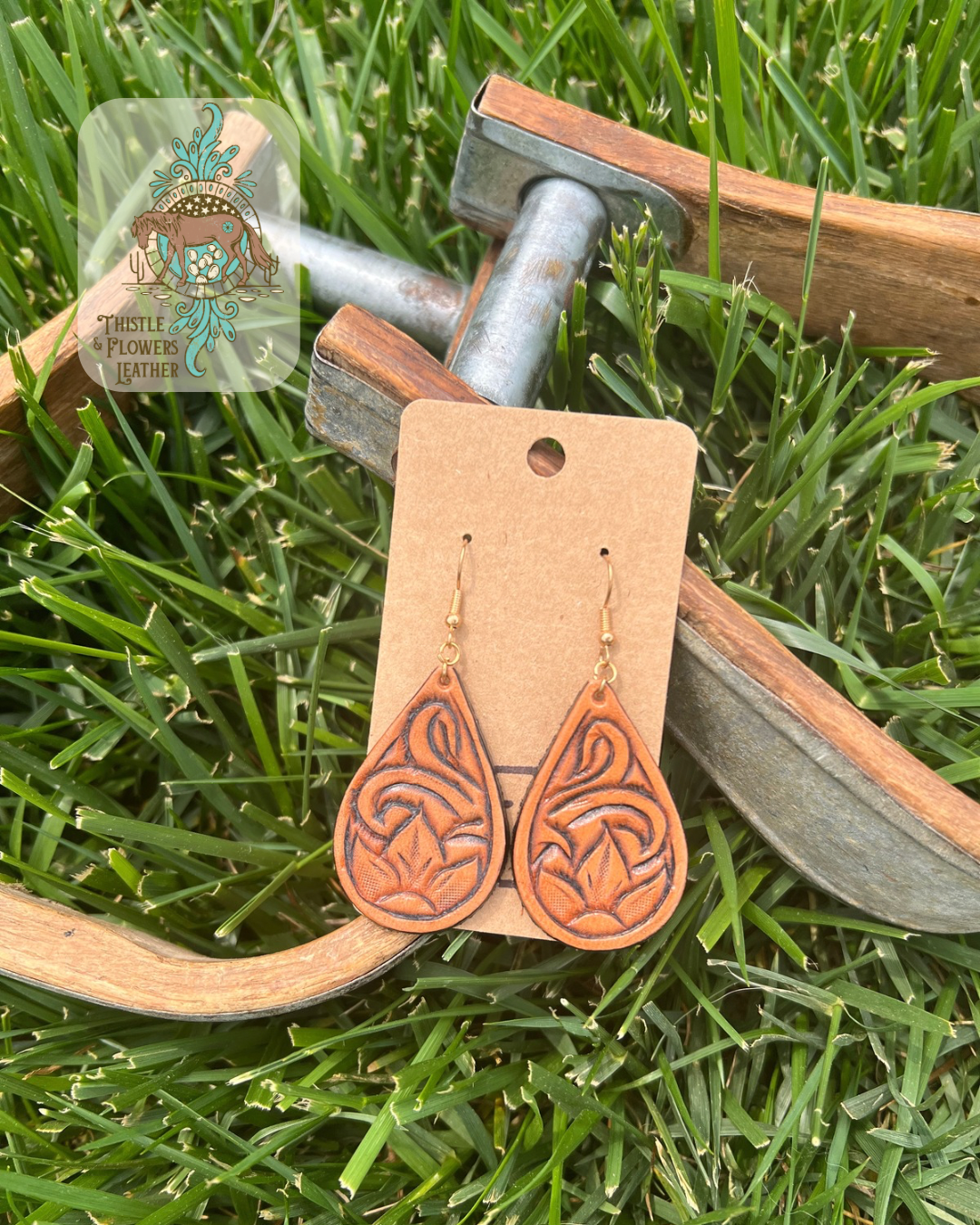 Teardrop shaped tooled leather earrings with a flower and traditional western tooling. Photographed on metal and wooden stirrups on grass, with logo on top left corner of photo.