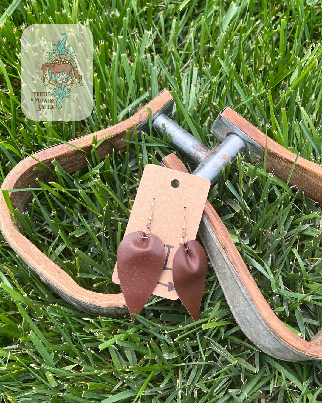 Smooth dark brown reverse teardrop leather earrings. Photographed on wooden and metal stirrups on grass with logo on top left corner of photo.