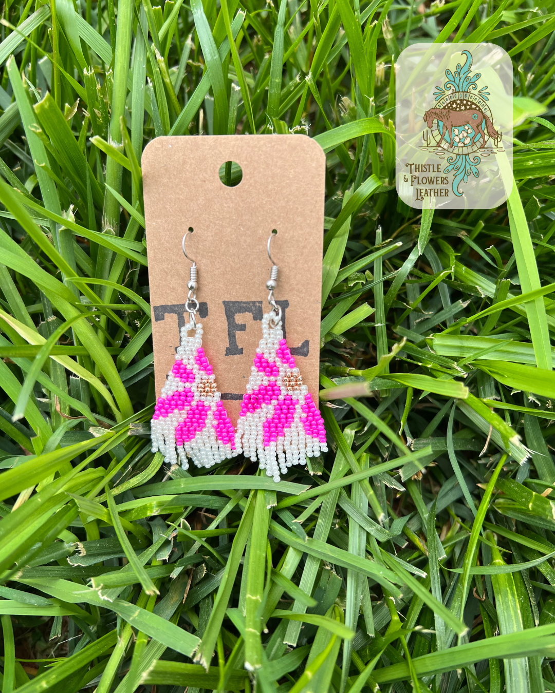 Hand beaded earrings with a daisy design. Bead colors are white, metallic gold, and hot pink. Photographed on grass, with logo on top right of photo.