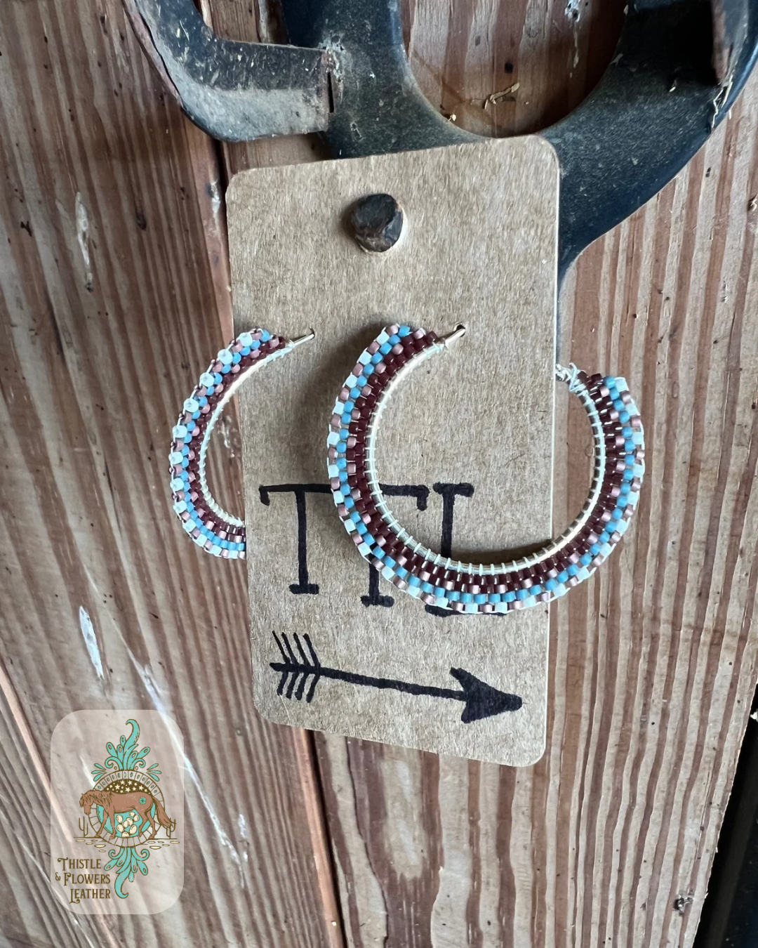 Beaded hoop earrings on silver colored base. Bead colors are copper, turquoise, brown, and cream. Beads make a pattern reminiscent of a western saddle pad. Earrings are hanging on a stall front. Logo is on bottom left corner of photo.