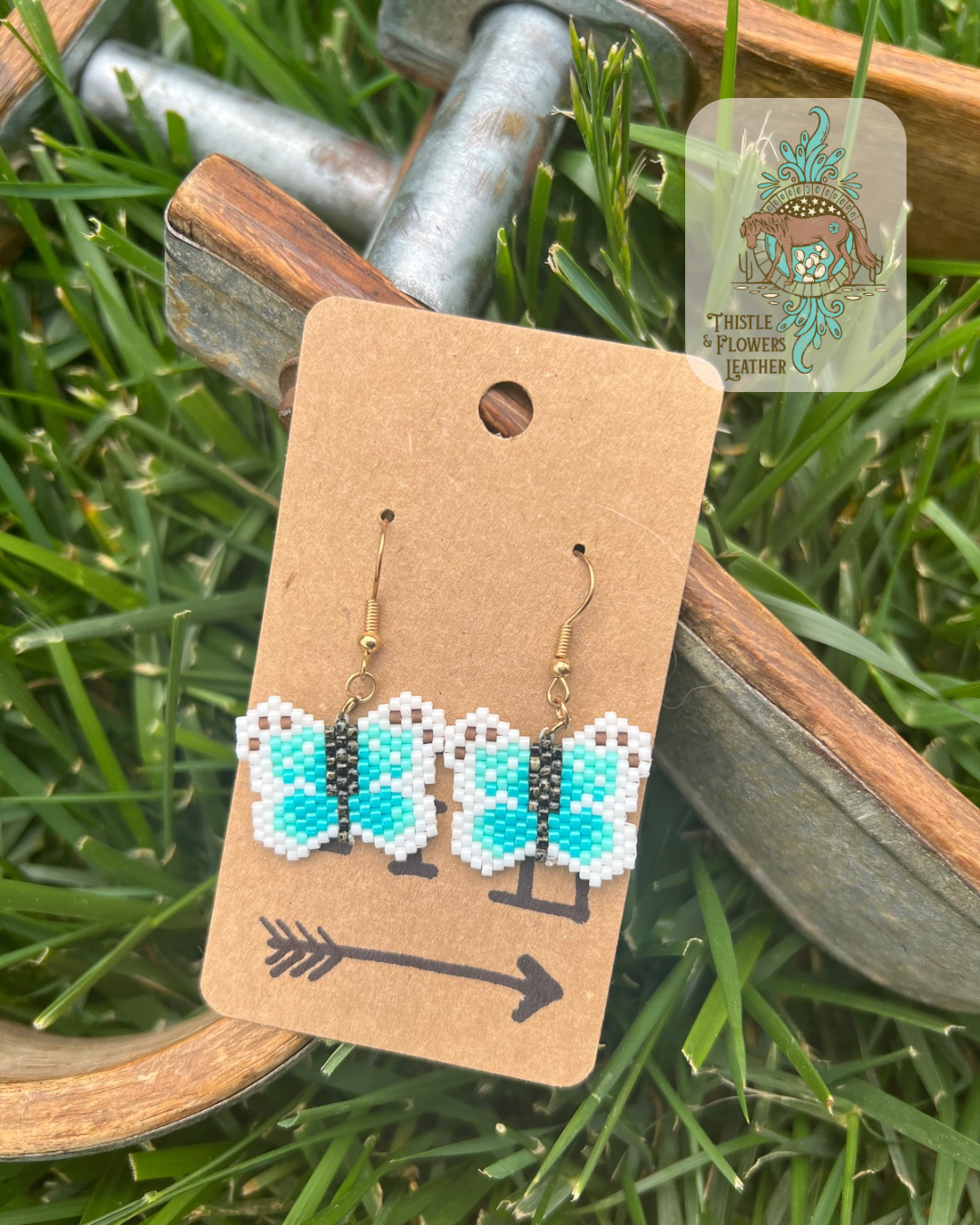 Mini monarch styled butterfly beaded earrings with beads in black, white, brown, and various shades of blue. Photographed on metal and wooden stirrups on grass with logo on upper right of photo.