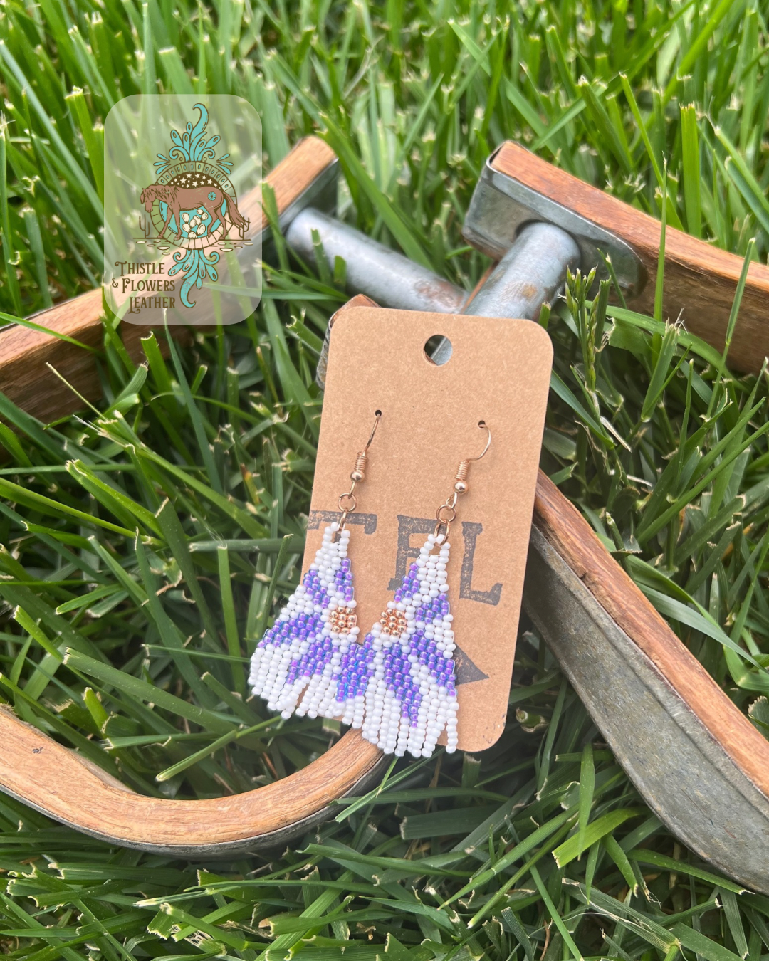 Beaded earrings with a daisy design. Bead colors a lighter purple, metallic gold, and white. Photographed on wooden and metal stirrups on grass, with logo on top left of photo.