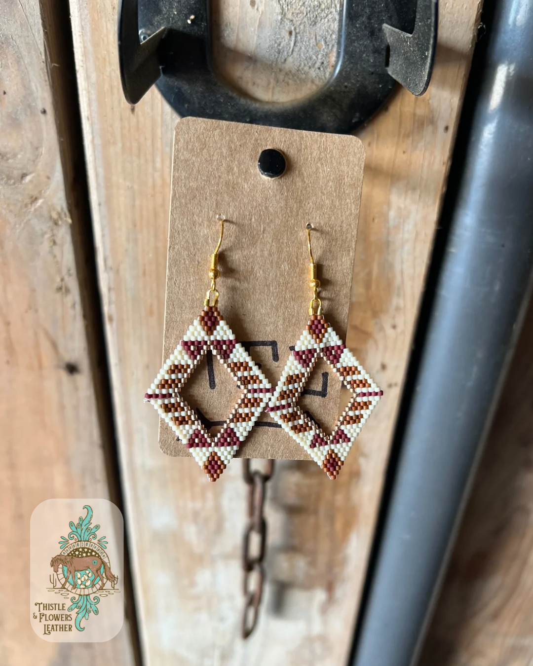 Beaded earrings. Beaded earrings with bead colors in cream, rust, brick red, and gold make up a diamond shape with a hollow diamond shaped center. Western design in beads. Earrings are hanging on a stall front. Logo is in bottom left corner of photo.