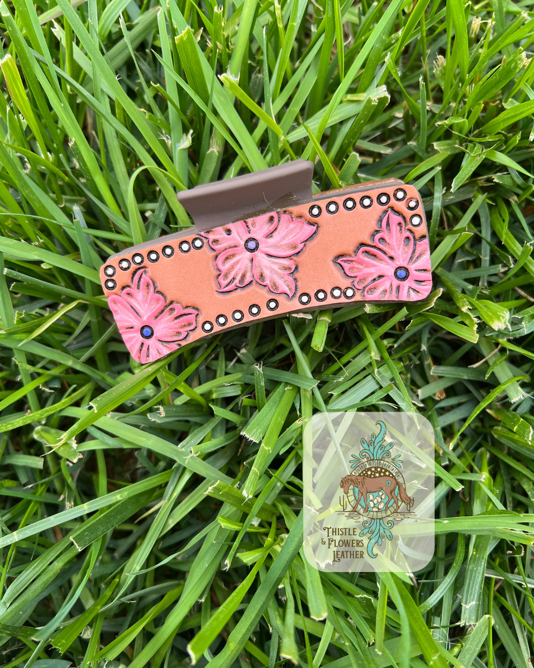 Brown Hairclip base with 3 tooled and painted pink flowers with purple center dots. White dotted tooled and painted border around leather patch. Photographed on grass with logo on bottom right of photo.