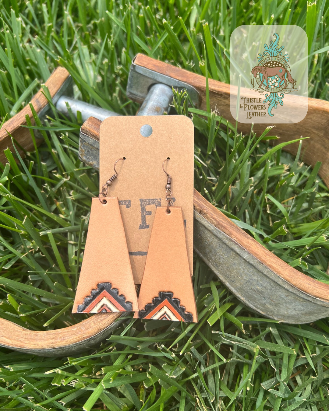 Long trapezoid shaped tooled leather earrings with a western inspired design on the bottom, tooled and painted black, rust, and white. Photographed against metal and wooden stirrups on grass. Logo is in top right corner of photo.