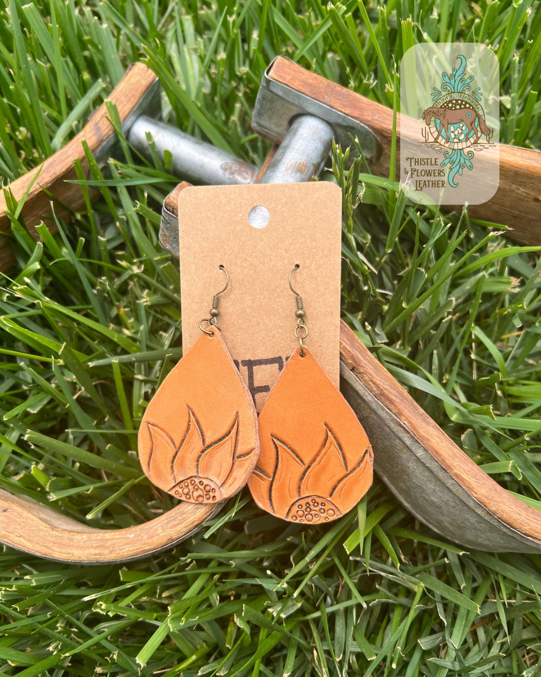 Teardrop shaped tooled leather earrings with tooled sunflowers on the bottom. Photographed on metal and wooden stirrups on grass with logo on upper right of photo.
