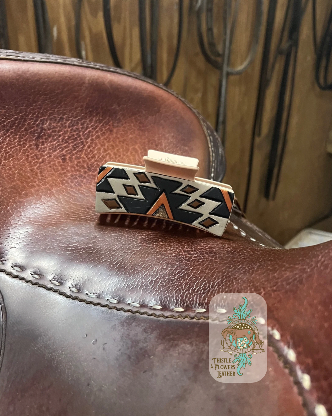 Large hair clip with cream colored base and a hand tooled and painted patch. Patch has a southwest design tooled and painted. Colors are gray, black, rust, and brown. Clip is on a saddle seat. Logo is on the top right of photo.