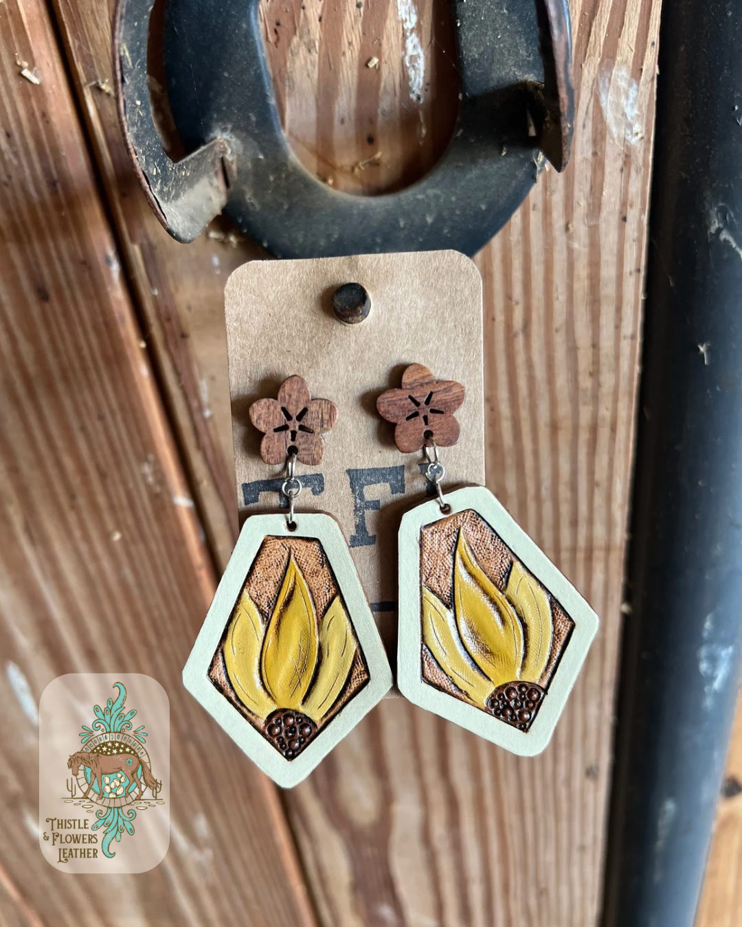 Hand tooled and painted leather earrings. Earrings have a wooden stud front carved in the shape of a flower (actual stud-in-ear part is not wooden) and a offset diamond shape tooled leather earring with a white border and a yellow sunflower with brown center at the top of the diamond. Earrings are hanging on a stall front. Logo is in bottom left of photo.