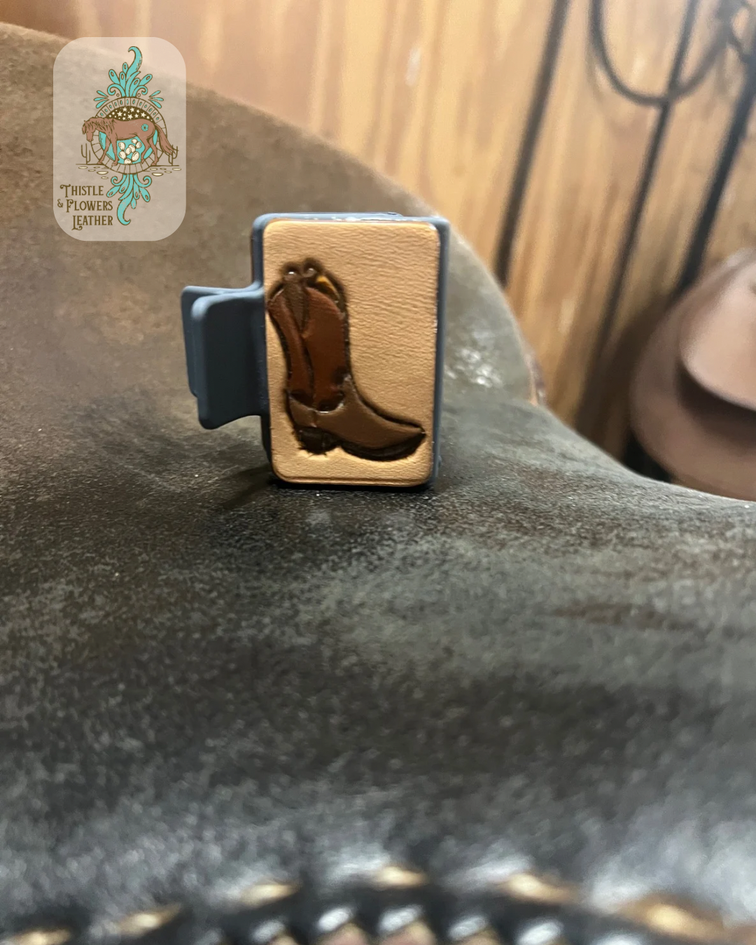 Blue base hairclip with a hand tooled and painted leather patch. Patch has a cowboy boot tooled and painted on it. Boot is painted various shades of brown. Clip is sitting on a saddle seat. Logo is in the top left corner.