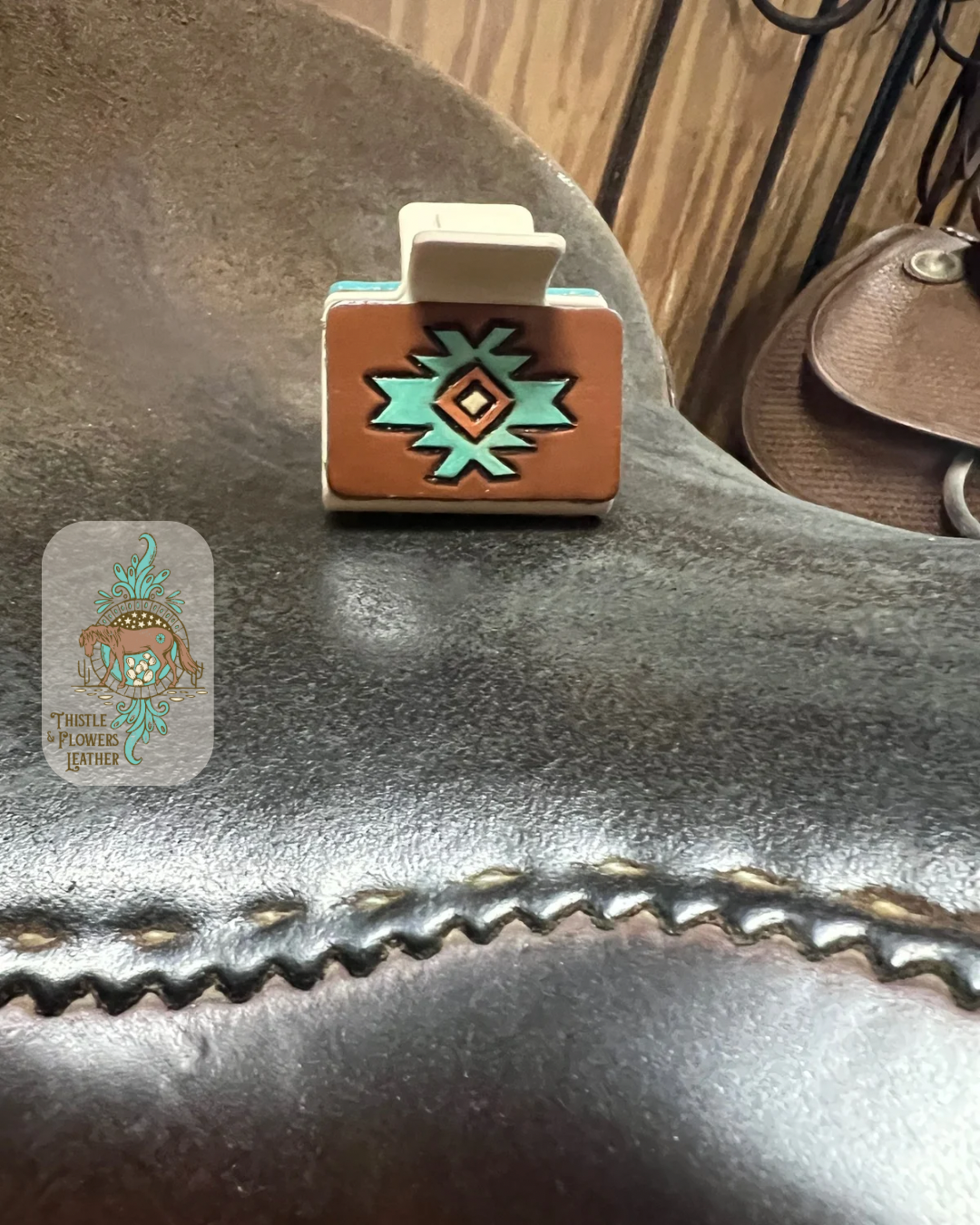 Medium cream base hairclip with hand tooled and painted leather patch. Patch is tooled with a southwestern design and the paint colors are brown, cream, turquoise, and metallic copper. Hairclip is resting on saddle seat. Logo is in the left side of photo.