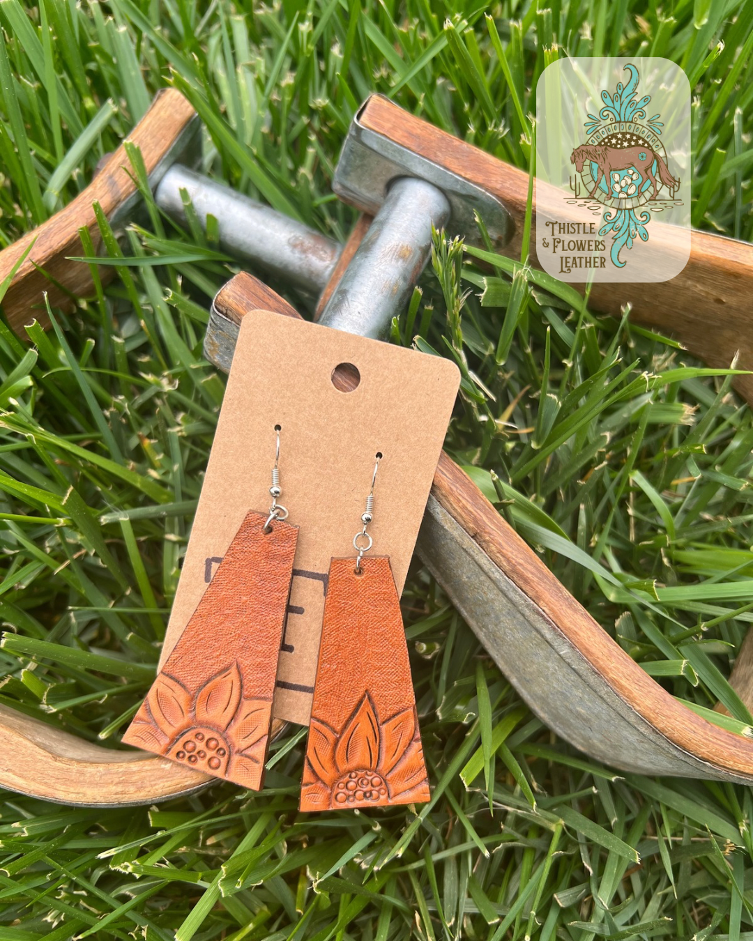 Tooled leather earrings in a long trapezoid shape with a sunflower on the bottom of each and backgrounding on the rest of the earring. Photographed on metal and wooden stirrups on grass with logo on top right of photo.