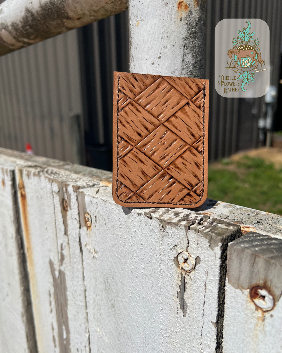 Handmade tooled leather cardholder with a large basketweave pattern. Photo shows the back of cardholder. Cardholder is leaning against an arena fence pole. Logo is in the top right corner of photo.