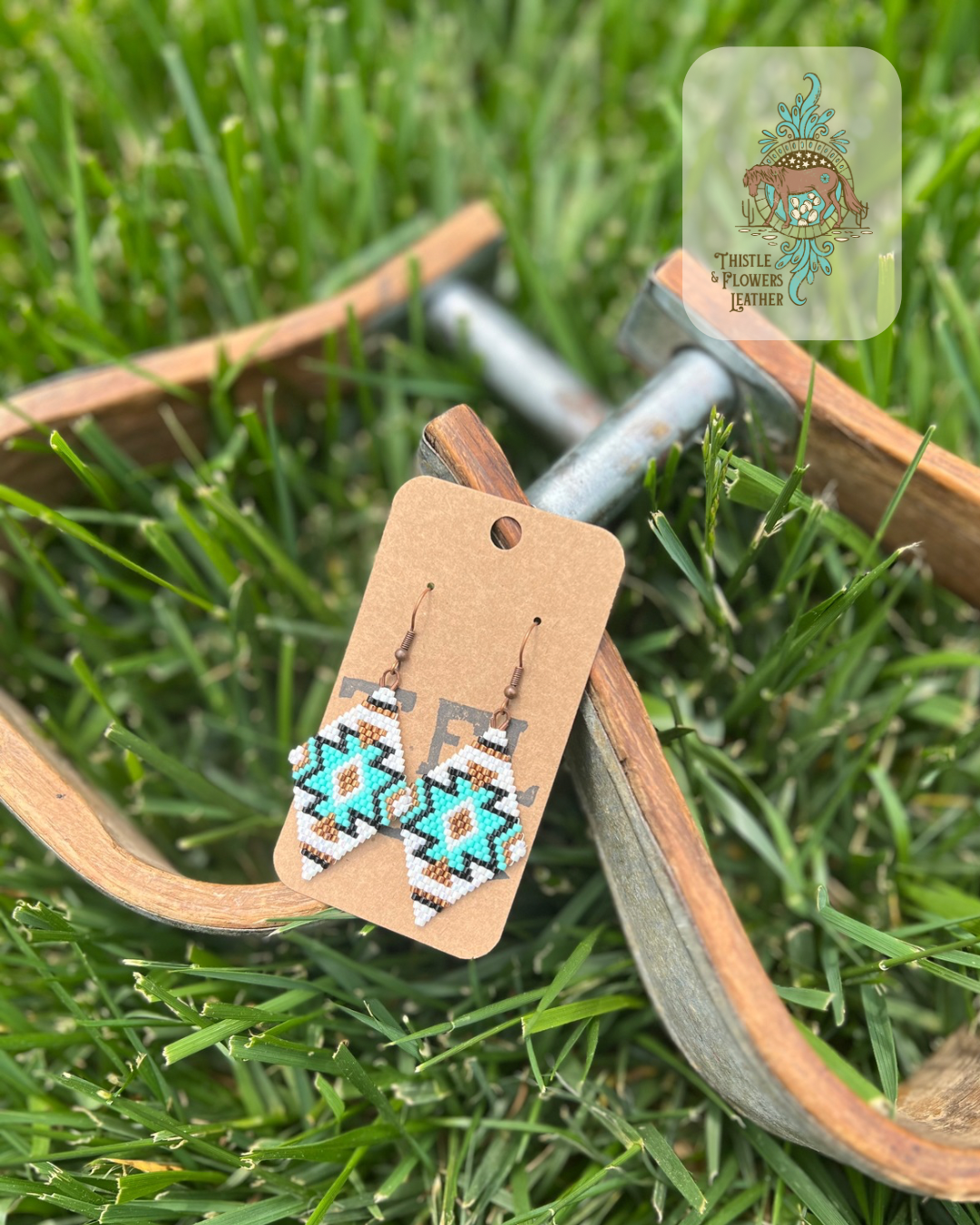Diamond shaped beaded earrings with a southwestern design. Bead colors are white, metallic gold, turquoise, and black. Photographed on metal and wooden stirrups on grass with logo on top right of photo.