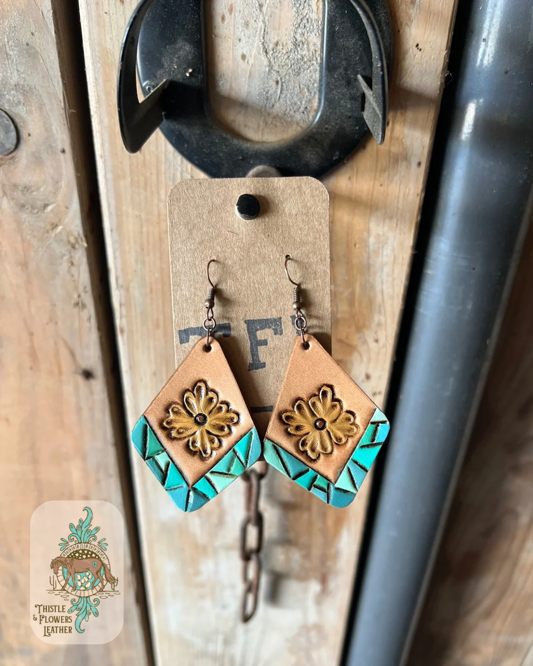 Tooled and painted leather diamond shaped earrings. Bottom edges have a tooled faux turquoise border and there is a yellow traditional western flower in the center. Earrings are hanging on stall front. Logo is the bottom left corner.