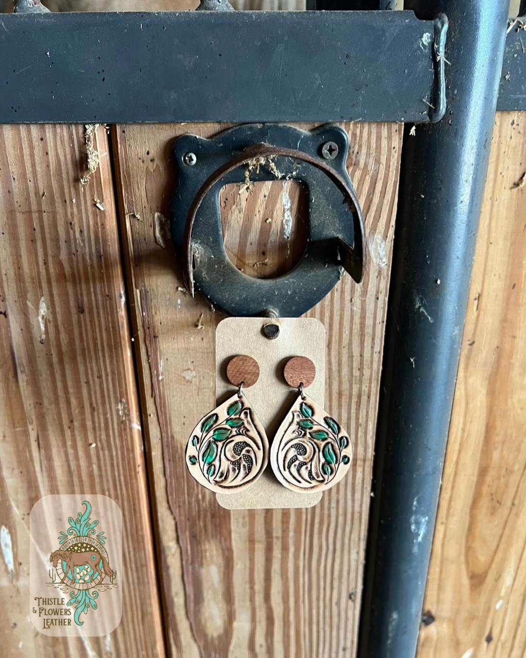 Hand tooled and painted leather earrings. Earrings feature a tooled traditional western design and a tooled and painted vine with leaves. Earrings are a teardrop shape dangling from a wooden circular stud front(actual stud-in-ear part is not wooden. Earrings are hanging from a stall front. Logo is in bottom left of photo.