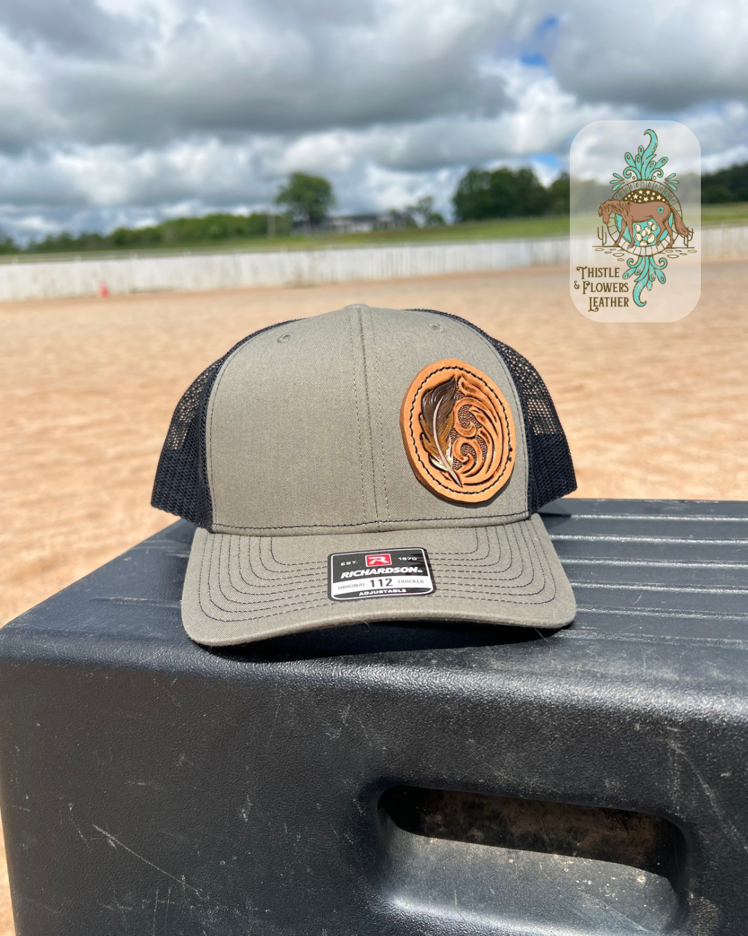 Tan baseball hat with black mesh and tooled leather circular patch. Leather patch has traditional Western tooling as well and a tooled and painted Feather. Patch is hand stitched on hat. Hat is on a mounting block in an arena. Logo is in top left of photo.