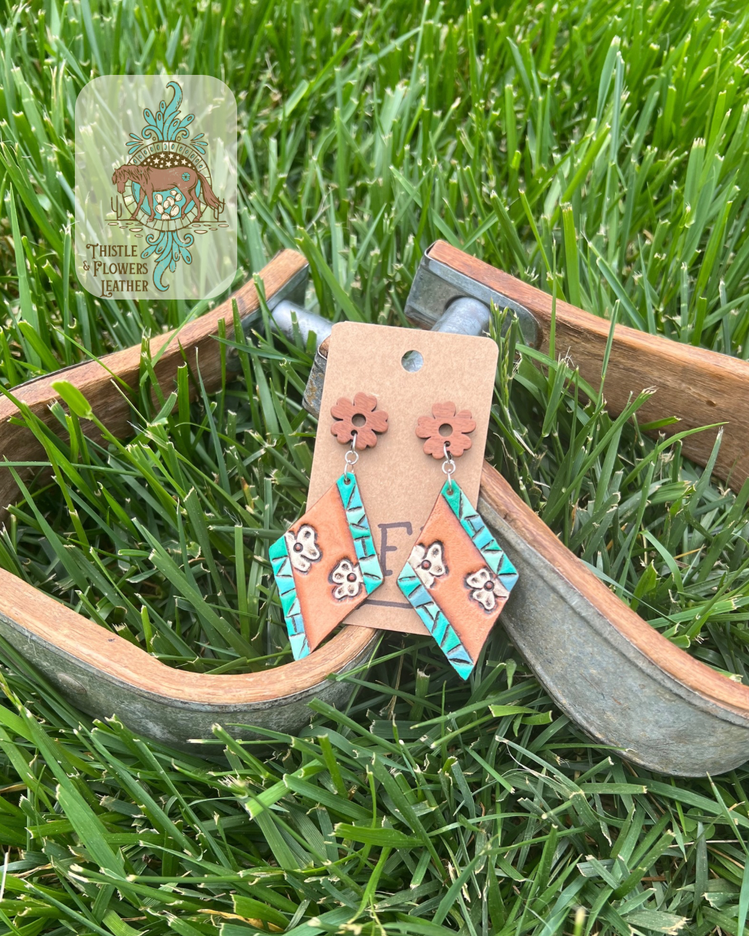 Tooled and painted leather earrings in a diamond shape with a wooden floral stud( actual stud piece is not wooden). Tooled design has white small flowers and 2 edges on each earrings are a faux turquoise design with different shades of turquoise and teal. Photographed on metal and wooden stirrups on grass with logo in the top left corner of photo.
