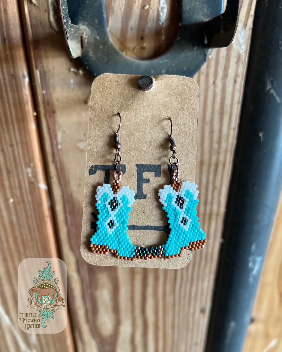 Hand beaded earrings in the shape of a cowboy boot. Bead colors are brown, white, copper, turquoise, and dark teal. Earrings are hanging on a stall front. Logo is in the bottom left of photo.