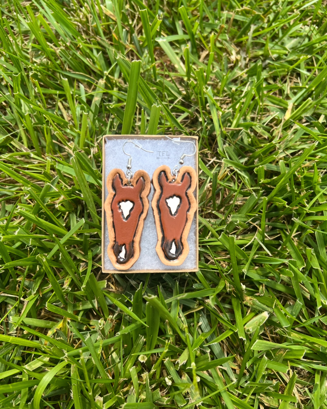 Custom tooled leather earrings designed to look like a bay horse in an earring box outside on grass. Earrings have horse head shape, natural leather colored border, and has markings exactly like the horse.