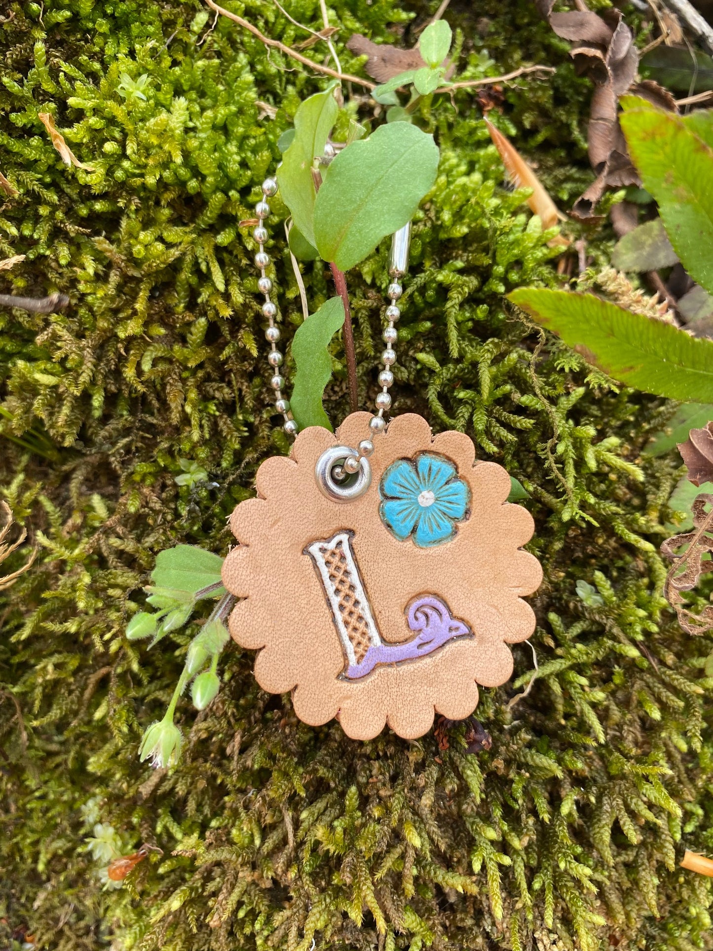 Scalloped edge leather charm with a tooled and painted letter "L" with a blue flower on a moss background. Silver keychain