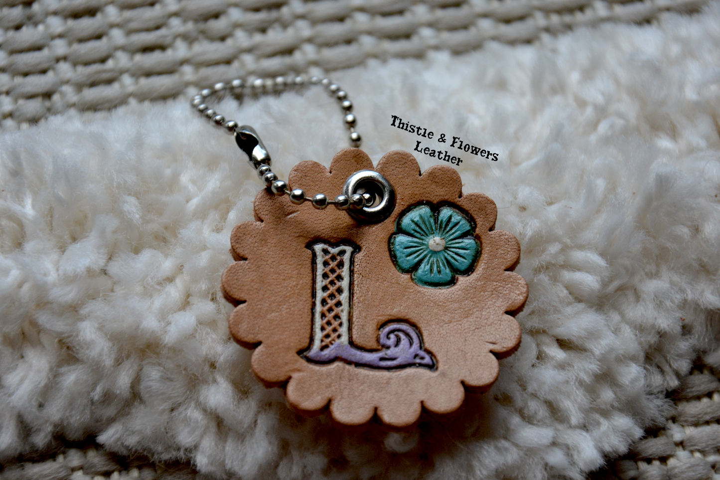 Tooled leather keychain with beaded connector. Scalloped edges, letter "L" with white and purple coloration. Blue and white flower. Antiqued.