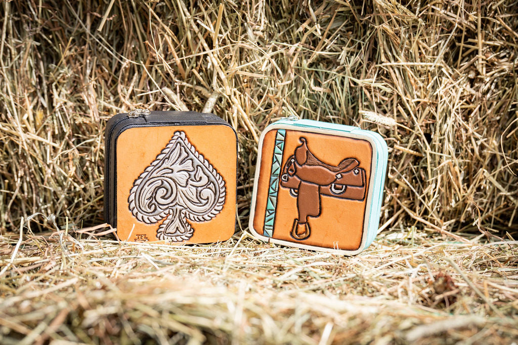 2 travel jewelry boxes with tooled & painted leather toppers (one with a faux engraved silver spade, one with a western saddle) both on a hay bale.