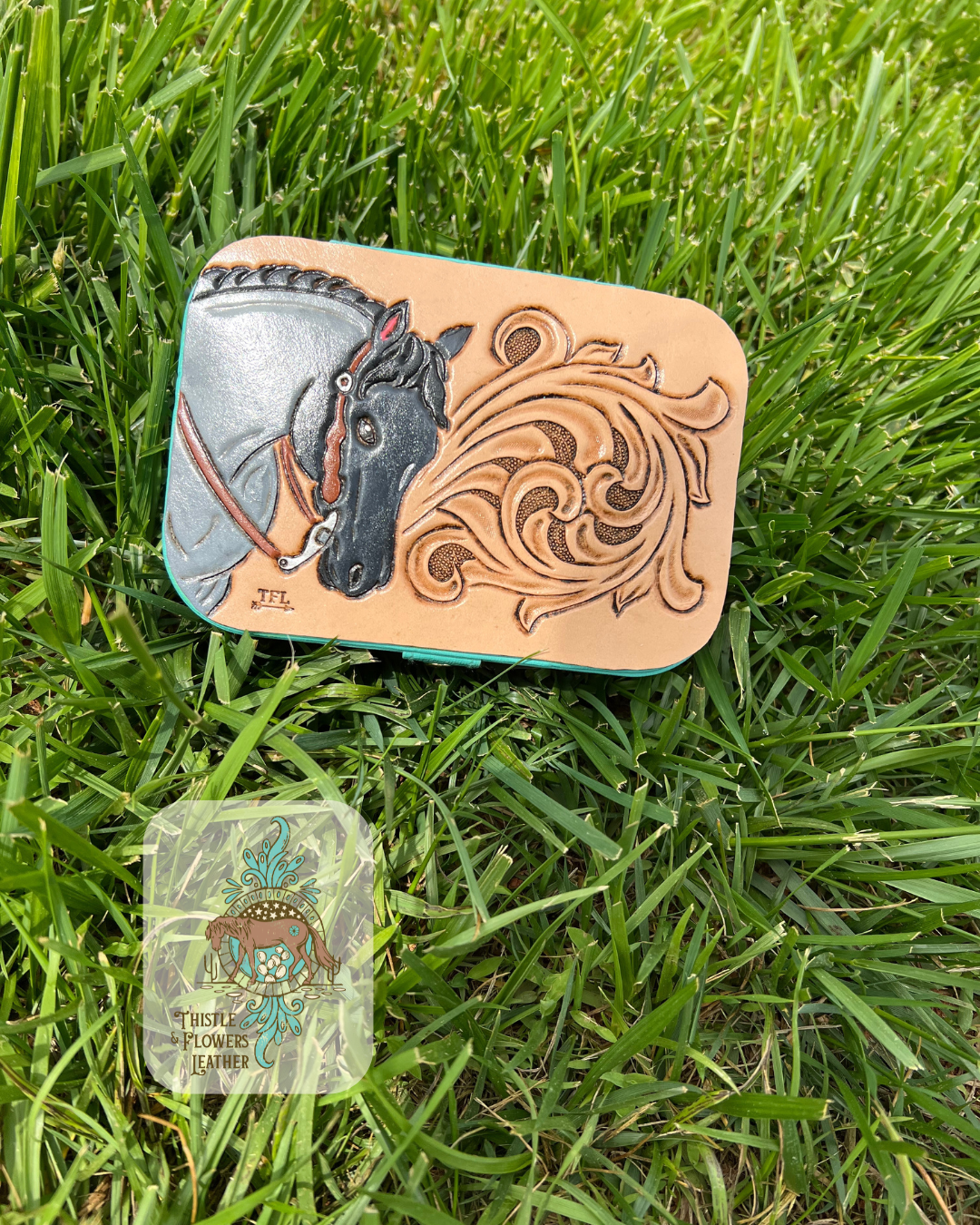 Medium sized jewelry box with a traditional western tooling design and a blue roan horse on the top. Jewelry box is on grass. Logo is on the bottom left.