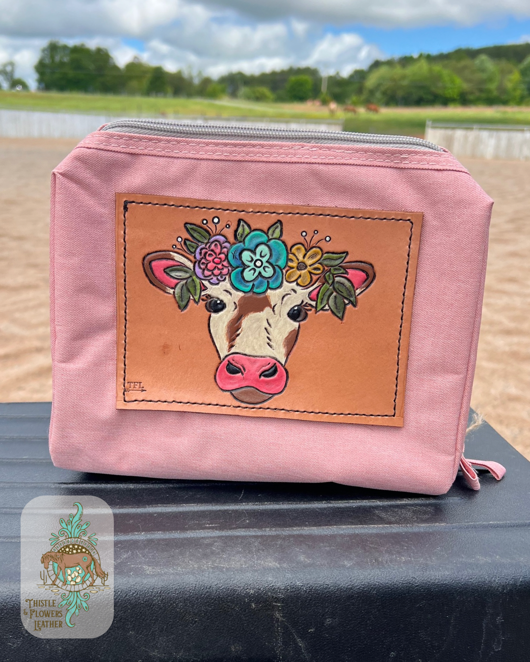 Pink makeup bag with tooled and painted cow in a flower crown. Makup bag is sitting on a mounting block in arena. Logo is in bottom left of photo.
