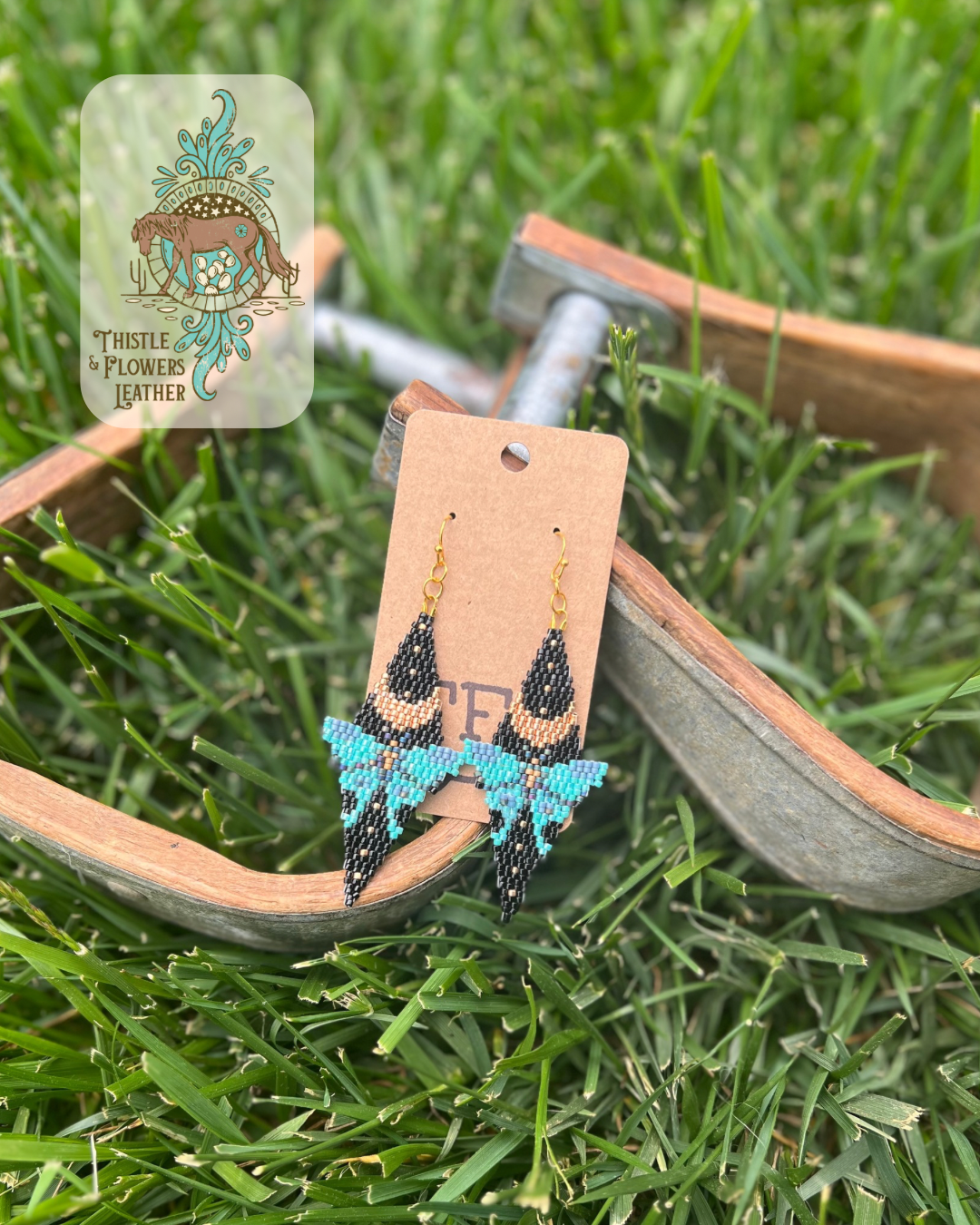 Diamond shaped lunar moth earrings, our "Chlo" style. Features black, gold, and shades of blue/turquoise beads. Earrings are laying on metal and wooden stirrups on grass. Logo is in top left of photo.