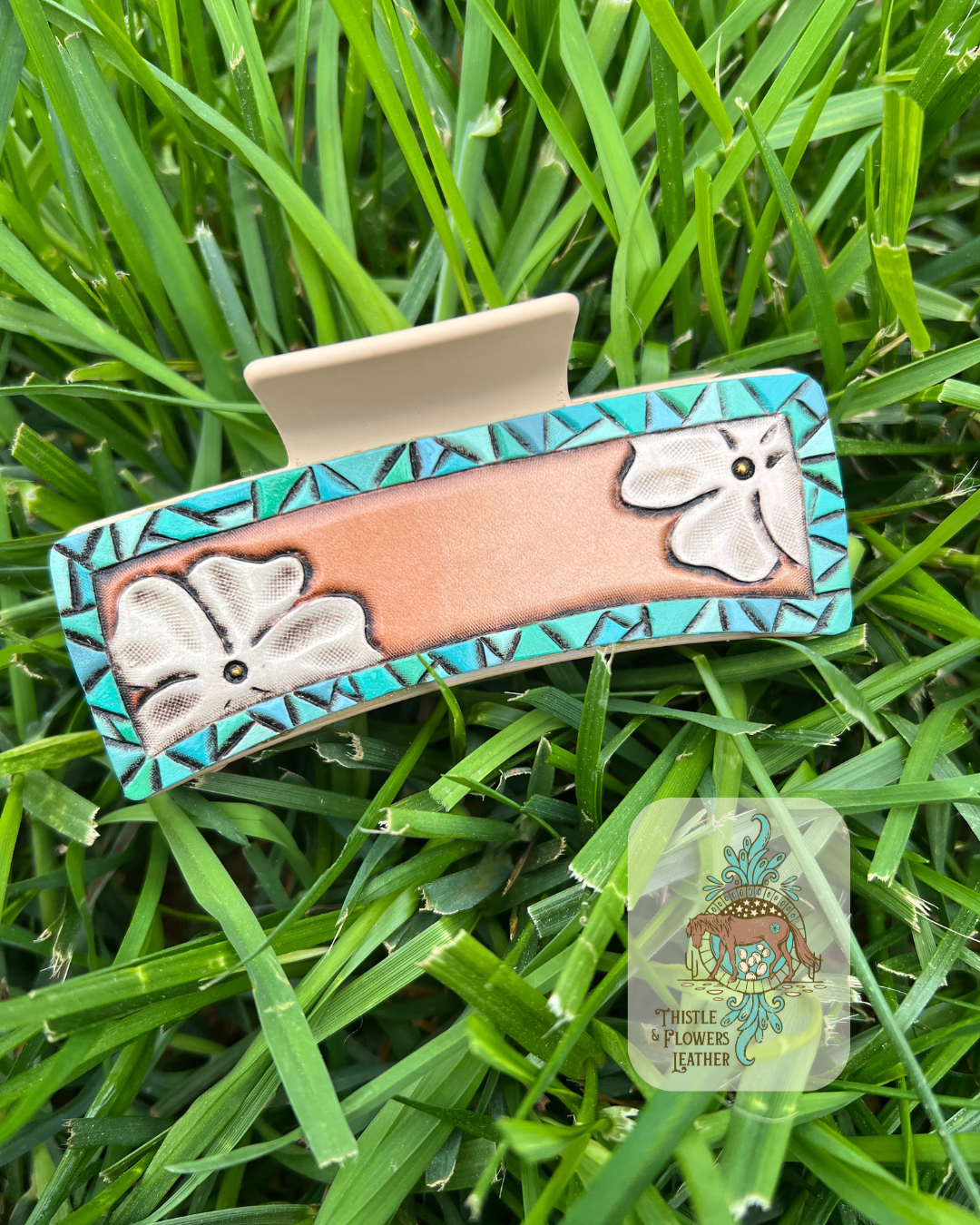 Large hairclip with cream colored base and hand tooled and painted leather patches. Patches have a faux turquoise tooled border and white flowers in opposite corners. Clip is sitting on grass. Logo is in the bottom right of photo.