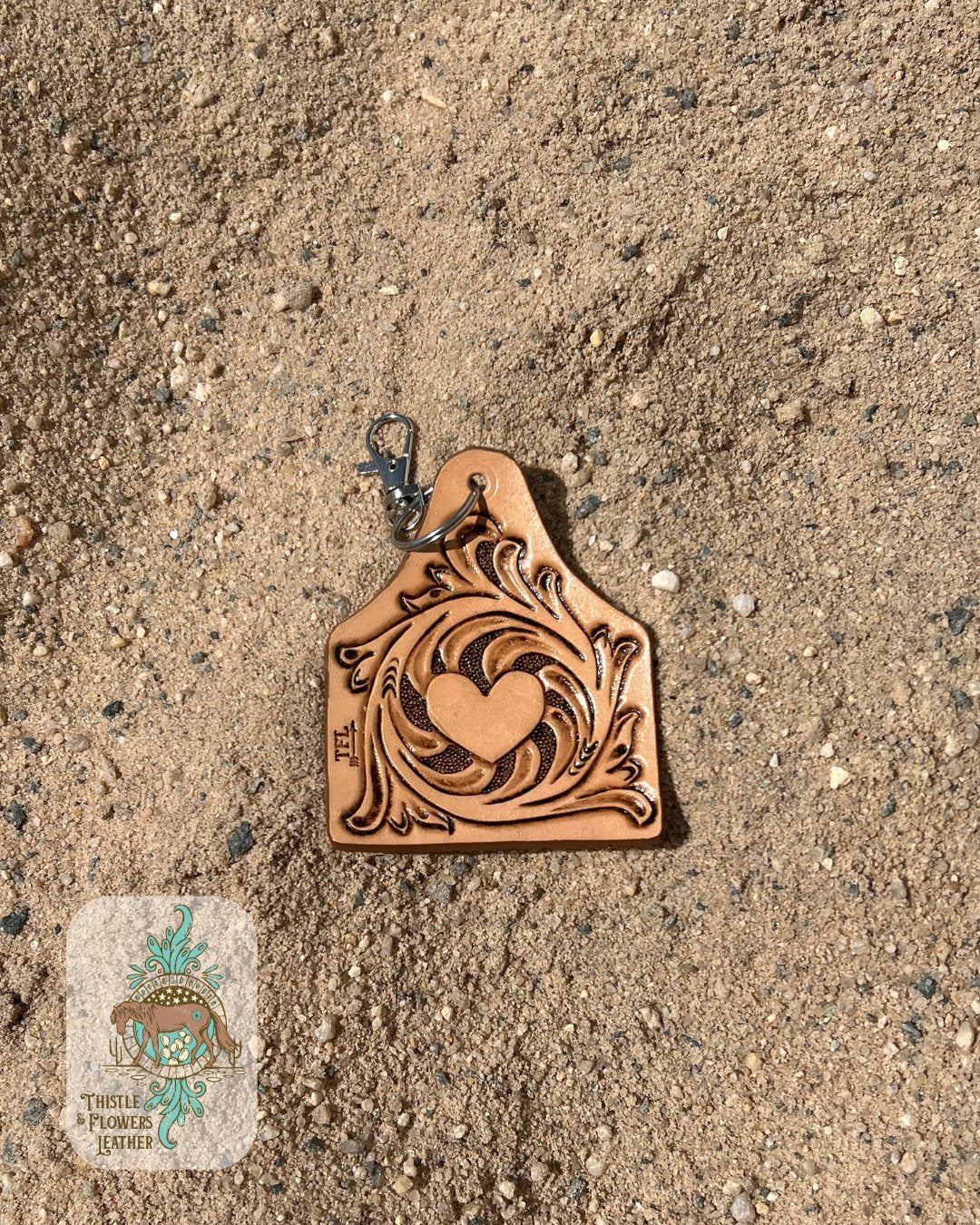 Cowtag shape & size tooled leather keychain. Features traditional tooling and a heart design in the center. Keychain is on arena sand. Logo is in bottom left corner of photo.