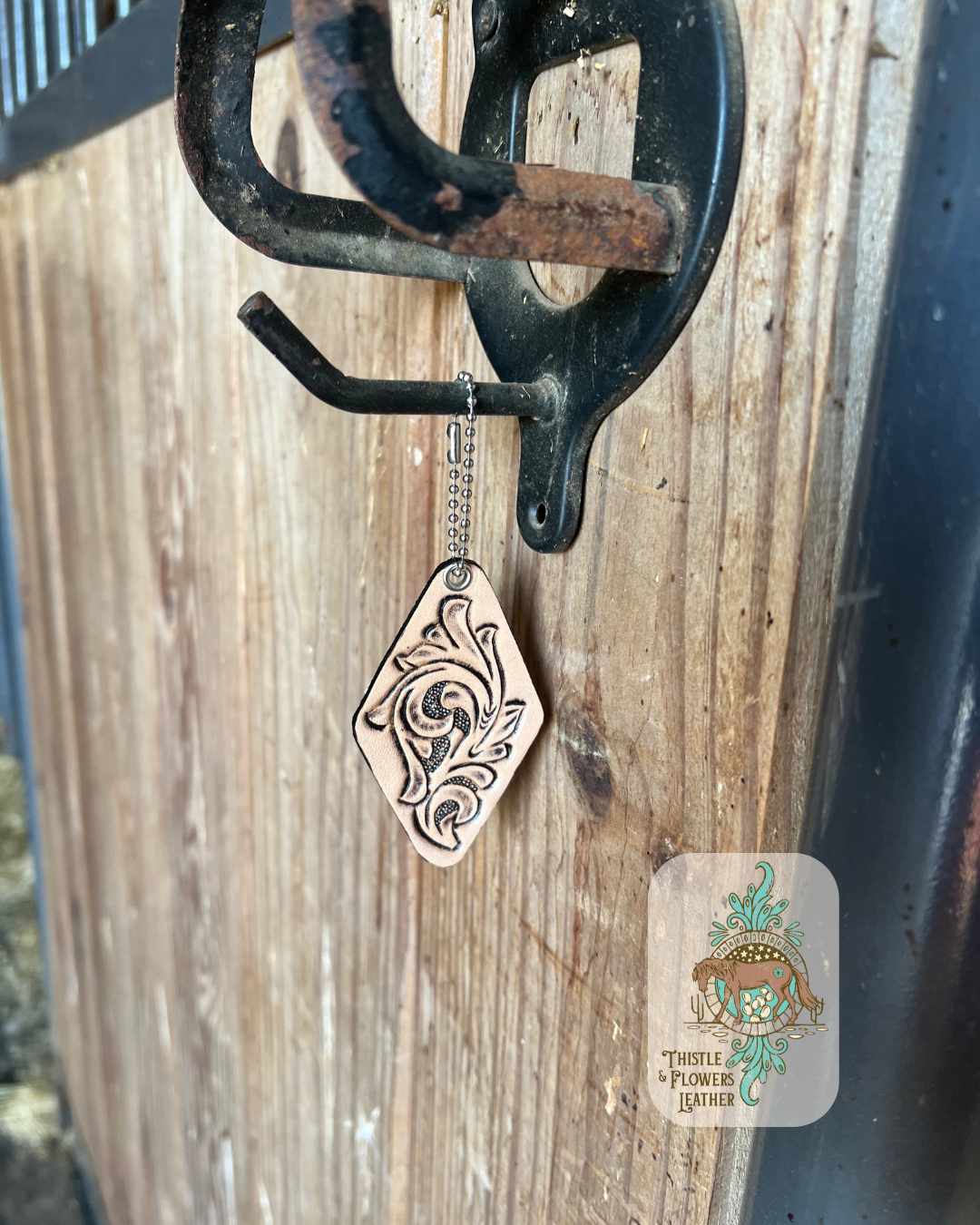 Hand tooled leather hotel style keychain with a traditional western tooling design. Keychain is hanging on stall front. Logo is in bottom right of photo.