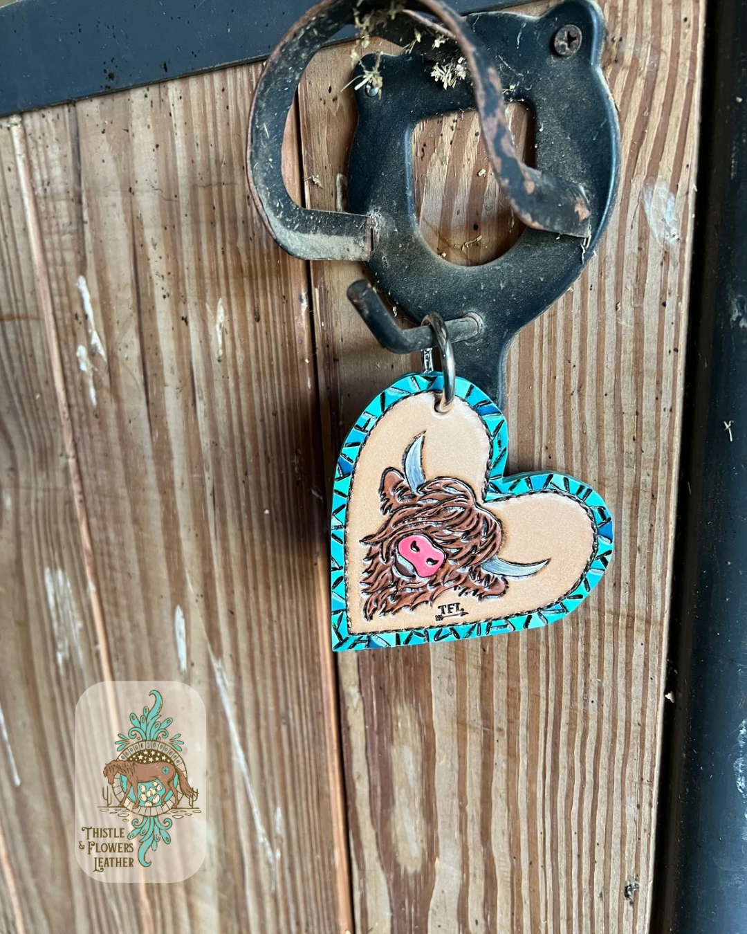 Heart shaped hand tooled and painted purse charm. Charm has a faux turquoise tooled border and a brown highland cow in the center. Hanging on a stall front. Logo is on bottom left of photo.