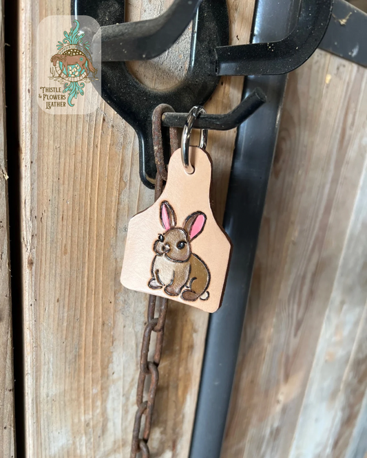 Tooled leather keychain in a goat tag size with a tooled and painted brown bunny with pink ears. Keychain is hanging on stall front. Logo is on top left of photo.