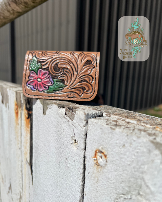 Cardholder with traditional western tooling and a flower with 2 leaves. Cardholder is hand painted and tooled. Leaves are green and flower is pink and purple. Cardholder is on arena fence. Logo is on top right of photo.