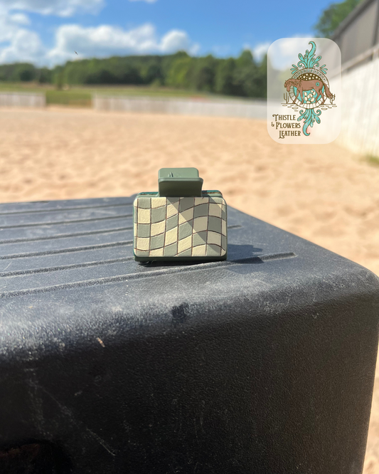 Green hair clip base with a cream and sage green wavy checkerboard pattern leather patch. Leather patch is hand tooled and painted. Hairclip is sitting on a mounting block in a riding arena. Logo is in top right of photo.