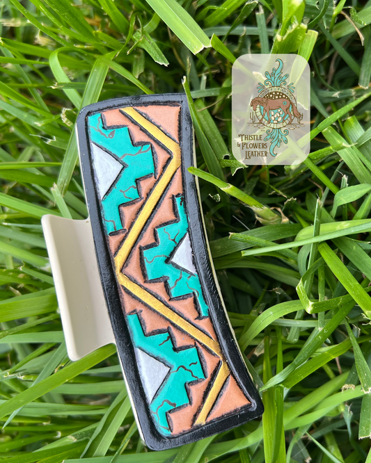 Cream base hair clip with Aztec designs painted a realistic faux turquoise with opalescent center, black border around leather patch, and golden zigzag accent line between Aztec designs. Photographed on grass with logo on top right corner of photo.