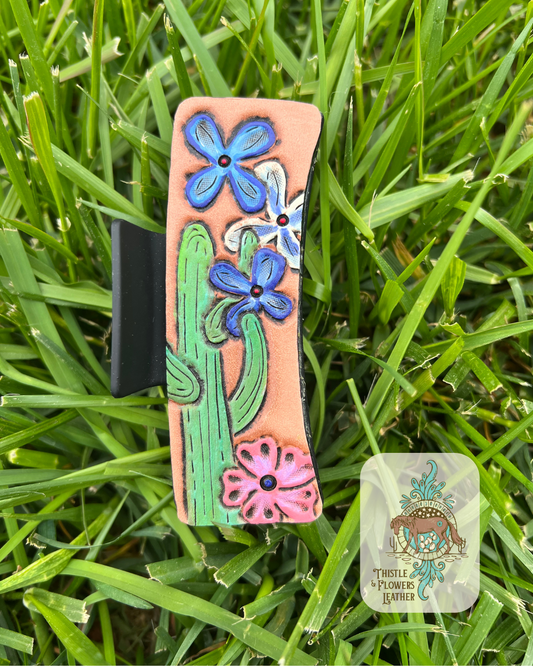 Black hair clip base with tooled & painted green cactus and 2 blue flowers, 1 white and blue flower, and 1 pink flower. Photographed on grass with logo in bottom right of photo.