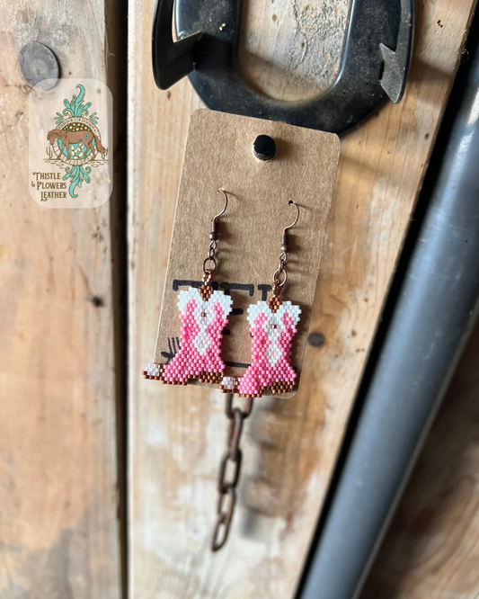Hand beaded earrings in the shape of a cowboy boot. Bead colors are brown, white, copper, pink, and light pink. Earrings are hanging on a stall front. Logo is in the top left of photo.