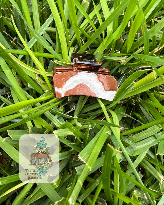 Clear ish brown hair clip with 2 tooled and painted leather patches. Patches are tooled and painted to look like brown cow print. Photographed on grass with logo in bottom left corner.